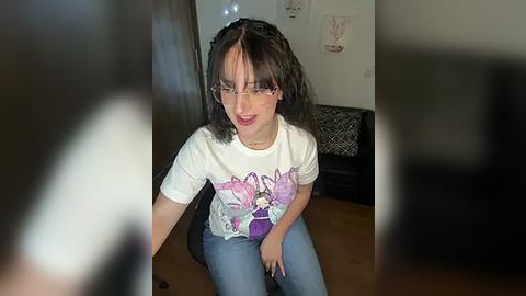 Media: Video of a young girl with glasses, short dark hair, wearing a white T-shirt with a purple butterfly graphic, sitting on a wooden floor in a cluttered room with artwork and furniture.