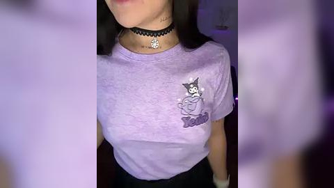 Media: A video of a young woman with light skin, wearing a lavender t-shirt with a cartoon character, black choker, and black pants, indoors.