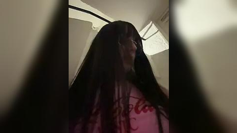 Media: A video showing a person with long black hair and a red shirt, partially blurred and seen from a low angle, in a dimly lit room with white walls and a skylight.