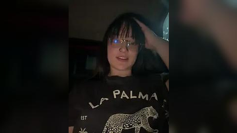 Media: Video of a woman with long dark hair and glasses, wearing a black \"LA PALMA\" t-shirt, sitting in a dimly lit room, hand on her head, with a blurred background.