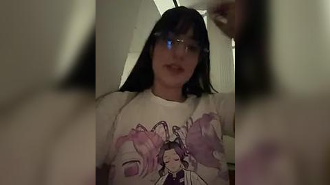 Media: Video of a young woman with dark hair, wearing glasses and a white T-shirt featuring a cartoon character, in a dimly lit room with blurred out faces.