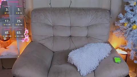 Media: Video of a cozy living room featuring a beige sectional couch with a white fuzzy throw pillow, lit by warm yellow lights. A decorated Christmas tree stands in the background, partially visible.