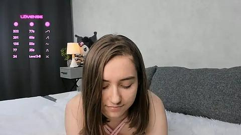 Media: A video of a young woman with shoulder-length brown hair, wearing a pink top, sitting on a bed with grey pillows. A stuffed animal and a digital display with stats are visible in the background.