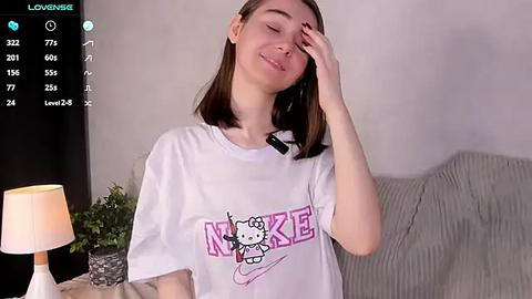 Media: Video of a young woman with straight brown hair, wearing a white T-shirt with a Hello Kitty design. She has fair skin and is touching her face. Background includes a beige couch and a green plant.