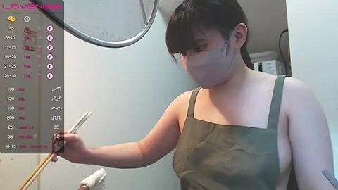 Media: A video of a young Asian woman with short black hair and glasses, wearing a green apron, using a hairbrush in a bathroom with a mirror and a \"LOVEN\" sign.