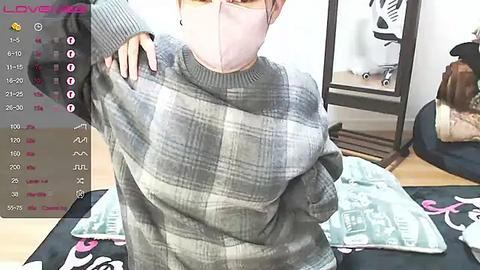 Media: Video of an Asian woman in a grey plaid sweater and mask, taken in a modern, brightly lit room with a plush green couch.