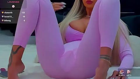 Media: Video of a blonde woman in a lavender bodysuit, lying on her back with legs spread, showcasing a tattoo on her right ankle. Background shows a modern indoor setting.