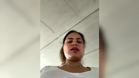 Media: Video of a young woman with light brown skin, brown eyes, and medium-length straight brown hair, wearing a white top. She stands against a white concrete ceiling, her mouth slightly open.