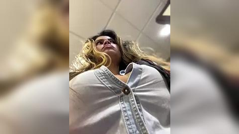 Media: Video of a young woman with long, wavy blonde hair, wearing a white denim jacket with a zipper, looking down from a high angle. Background shows a ceiling with fluorescent lights.