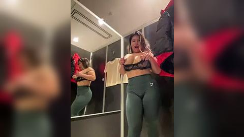 Media: Video of a curvy, dark-haired woman in a black lace bra and green leggings, standing in a dressing room mirror, pulling down her bra, revealing her large breasts.