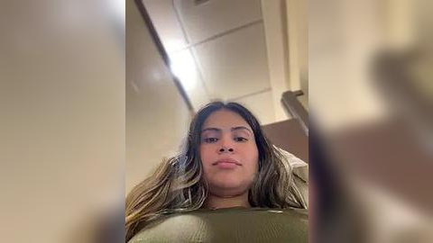 Media: A candid video of a young woman with long, wavy hair and light brown skin, wearing a green top, taken from a low angle in a dimly lit room with beige walls and a fluorescent ceiling light.