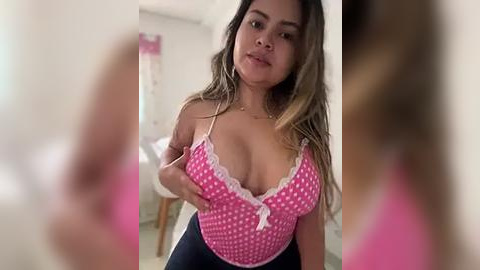 Media: Video of a Latina woman with long, wavy hair, wearing a pink polka-dot crop top, exposing ample cleavage, standing in a softly lit bedroom.