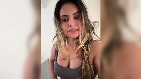 Media: Video of a Latina woman with long, wet, dark hair and tan skin, wearing a gray tank top that accentuates her large breasts, indoors with blurred background.