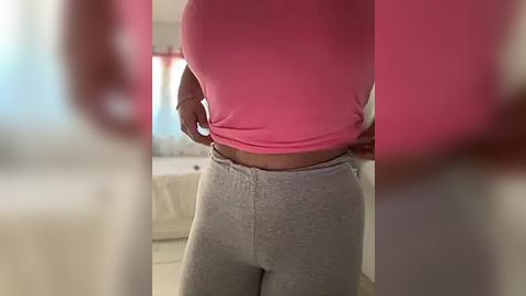 Media: Video of a woman in a bright pink top and gray sweatpants, standing in a softly lit room with beige walls and a white couch.