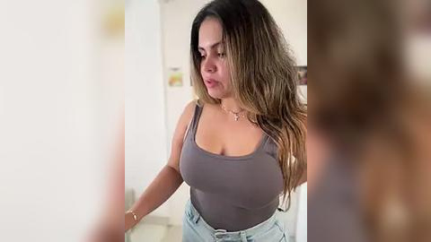 Media: Video of a curvy Latina woman with long, wavy brown hair, wearing a tight, grey tank top and high-waisted jeans, standing in a bright, indoor setting.