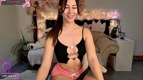 Media: Video of a smiling, light-skinned woman with long brown hair in a black cut-out crop top, pink shorts, and a belly button piercing, sitting in a cozy, decorated bedroom.