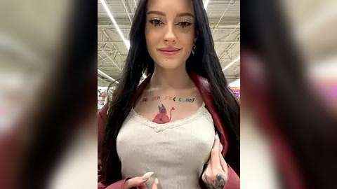 Media: Video of a young woman with long black hair, wearing a beige tank top, maroon jacket, and silver hoop earrings, with tattoos on her chest and forearm, standing in a mall under fluorescent lights.