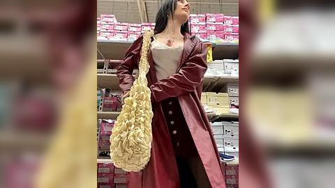 Media: Video of a woman in a red trench coat, white top, and black leggings, holding a beige floral handbag, standing in a cluttered, brightly lit retail store filled with pink and white cardboard boxes.