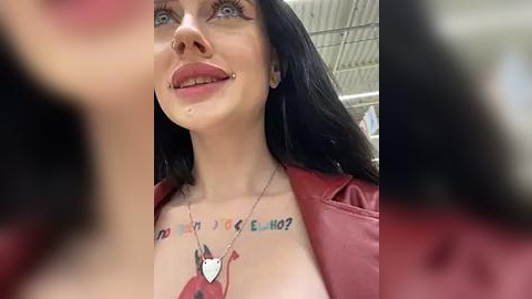 Media: Video of a smiling woman with pale skin, dark hair, and red lipstick, wearing a red jacket and a necklace. She has a tattoo across her chest reading \"Elviro.\