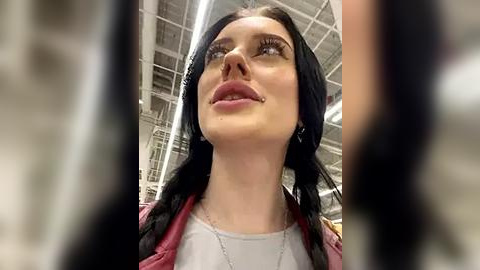 Media: Video of a young woman with long black hair, wearing a red jacket and a white top, looking upward in a warehouse setting with visible industrial ceiling beams.