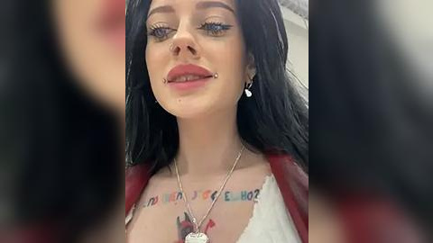 Media: Video of a young woman with pale skin, black hair, blue eyes, and a silver septum piercing. She wears a white lace bra, red jacket, and a necklace. Tattoo with \"WELCOME HOME\" across her chest. Background is blurred.