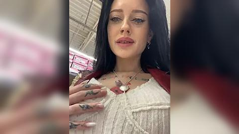 Media: A video of a young woman with long black hair, fair skin, and a delicate nose, wearing a white knit sweater and a heart pendant necklace, taken in a brightly lit room with a blurred background.