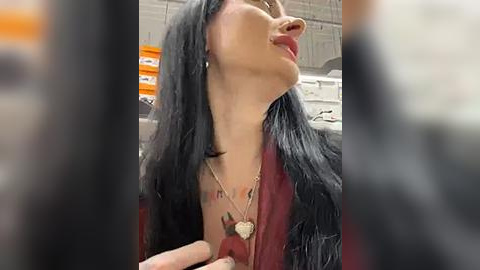 Media: Video of a woman with long black hair, fair skin, and a tattoo on her chest, wearing a black fur coat and a heart pendant necklace. Background shows a blurry office with white walls and an orange box.