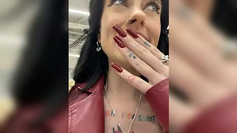 Media: Video of a woman with long black hair, red lipstick, and red nails, wearing a red leather jacket and multiple necklaces. She covers her mouth with her hand, revealing a tattoo on her wrist.