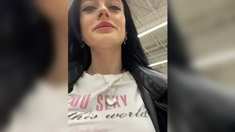 Media: Video of a young woman with long black hair, wearing a white T-shirt with \"YOU SEXY THING\" in pink and black text. She has a confident, slightly smirking expression, and is indoors with a blurred, industrial background.