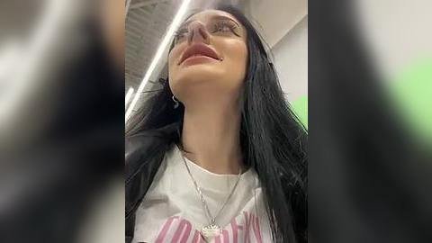 Media: Video of a young woman with long black hair, wearing glasses and a white T-shirt with pink text, looking upwards with a slight smile. Background includes a ceiling and blurred figures.