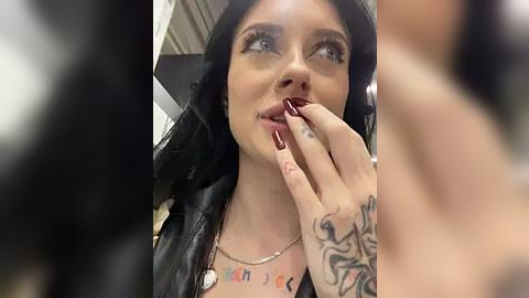 Media: A close-up video of a young woman with long black hair, wearing a black leather jacket and a gold necklace. She has a tattoo on her left hand and dark red nail polish, and she's licking her fingers suggestively.