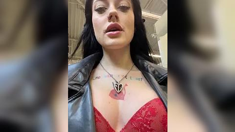 Media: A video of a fair-skinned woman with long black hair, wearing a red lace bra, black leather jacket, and a necklace with a heart pendant. Her lips are slightly parted, and she has tattoos on her chest.