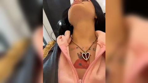 Media: Video of a woman with medium brown skin, wearing a pink jacket, heart necklace, and a tattoo of a red rose on her chest. Her black hair is partially visible.