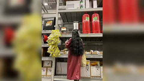 Media: Video of a supermarket aisle with a woman wearing a red dress, black hair, holding a yellow package, looking at red containers on shelves.