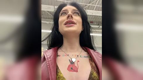 Media: Video of a woman with long black hair, fair skin, and a small tattoo of a red rose on her chest, wearing a pink leather jacket and a yellow shirt, looking upward.