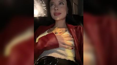 Media: A video shows a woman with pale skin, dark hair, and red lipstick, wearing a red jacket over a yellow top. She has a serene expression, holding a teddy bear, seated in a dimly lit room.