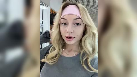 Media: Video of a blonde woman with fair skin, wearing a pink headband and gray sweater, posing indoors with blurred background.