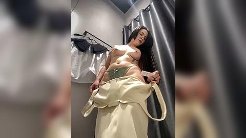 Media: Video of a woman with long dark hair, partially undressed in a dressing room, holding beige pants.