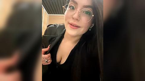 Media: Video of a young woman with fair skin, green eyes, and long dark hair, wearing large round glasses and a black top, smiling and holding a black bag in a dimly lit room.