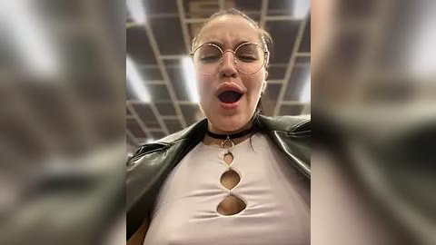 Media: Video of a woman with light skin and glasses, wearing a white top with cutouts and a black leather jacket, mouth open in surprise, in a dimly lit office with fluorescent lights.