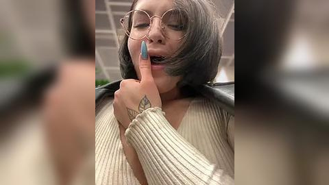 Media: Video of a young woman with shoulder-length, straight, dark brown hair and fair skin, wearing round glasses and a cream ribbed sweater. She's biting her nail with a blue manicure. Background is blurred.