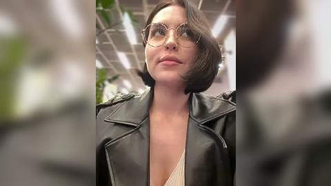 Media: Video of a woman with shoulder-length dark hair, wearing oversized round glasses and a black leather jacket over a low-cut top, standing in a modern, brightly lit room with green plants and industrial ceiling.