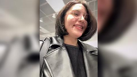 Media: Video of a smiling Asian woman with straight, shoulder-length dark hair, wearing glasses, a grey leather jacket over a black top, indoors with blurred background.