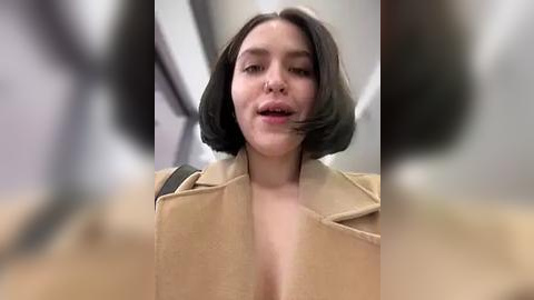 Media: A video of a woman with short black hair and fair skin wearing a beige coat, blurred background.