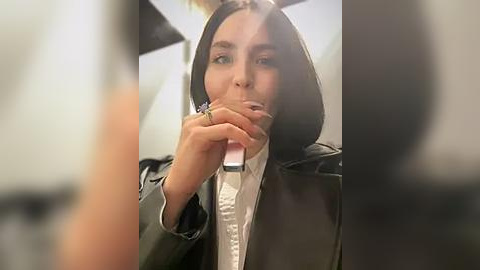 Media: Video of a woman with long black hair, wearing a white shirt and black blazer, holding a phone to her mouth, blurred figures in the background.