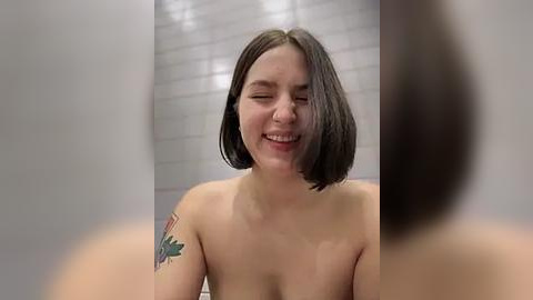 Media: Video of a topless young woman with fair skin and shoulder-length dark brown hair, smiling, in a tiled bathroom. Her left arm has a colorful tattoo.