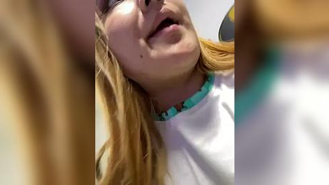 Media: Video of a young, smiling girl with long, wavy blonde hair, wearing a white shirt with turquoise trim. She has fair skin and is laughing, with a slight tilt of her head. The background is blurry.