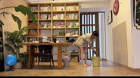 Media: Video of a woman in a beige dress and bare feet, cleaning the wooden floor with a yellow bucket. She stands in a well-lit room with a wooden desk, bookshelves, and plants.