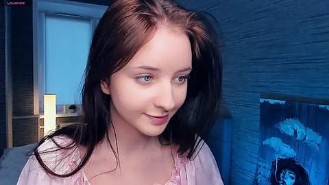 Media: Video of a young woman with fair skin, long brown hair, and blue eyes, wearing a light pink top, smiling softly, standing indoors with blue-toned walls and a decorative painting in the background.