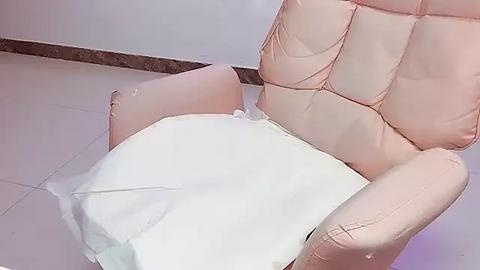 Media: Video of a person in a pink, quilted armchair with a white blanket covering their legs, sitting on a tiled floor with a brown marble baseboard. The person\u2019s face is not visible.